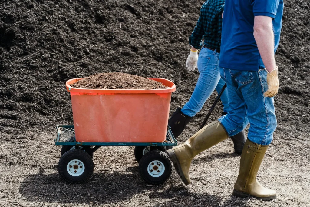 Person pulling a garden cart loaded with fresh compost past a large compost heap